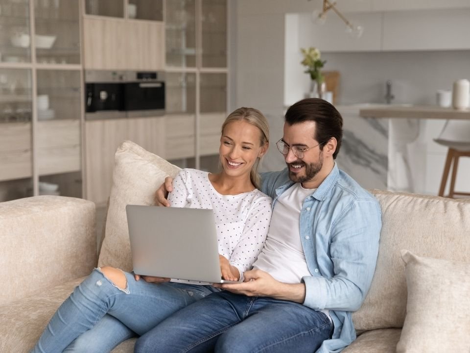 A couple at their kitchen table with a laptop, going over household numbers together and smiling.